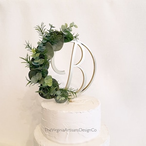 May include: A cake topper featuring the letter "B" in a silver and gold design, encircled by a wreath of green eucalyptus and other greenery. The topper sits atop a white frosted cake. The Virginia Artisans Design Co. is printed on the cake.