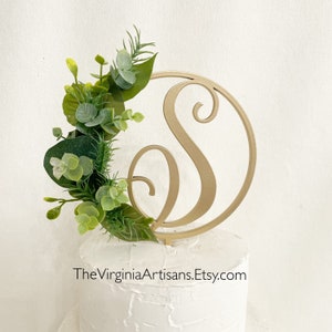 May include: A gold-colored cake topper featuring the letter "S" within a circle, adorned with green and light green faux foliage. The topper is placed on a white frosted cake. The text "TheVirginiaArtisans.Etsy.com" is visible.