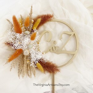 May include: A wedding cake topper featuring the initials "C & Z" in a gold-colored circular design. The topper is adorned with a bouquet of dried flowers in shades of orange, brown, and white. TheVirginiaArtisans.Etsy.com is printed at the bottom.
