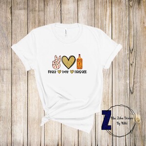 Pode incluir: Camiseta branca com um sinal de paz, um cora&ccedil;&atilde;o e uma garrafa de u&iacute;sque Fireball. O texto diz "Peace Love Fireball".