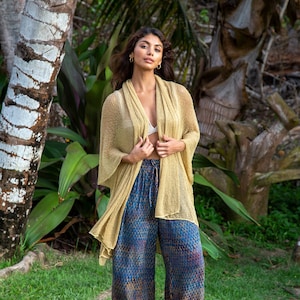May include: A woman wearing a light yellow sheer cardigan over blue and gold patterned trousers. She is standing in a tropical setting with lush green foliage.