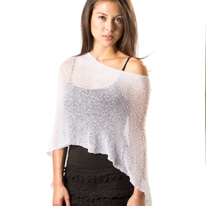 Pu&ograve; includere: Un poncho bianco in maglia larga e semitrasparente con orlo asimmetrico, indossato sopra una canotta nera e pantaloncini neri all'uncinetto con bordo smerlato. Il tessuto leggero a trama aperta presenta un sottile luccichio.
