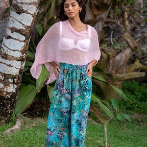 May include: A light pink, sheer, loose-knit poncho worn over a light pink bra, paired with wide-leg trousers featuring a vibrant, multi-coloured abstract pattern in shades of teal, green, blue, and pink with a drawstring waist.