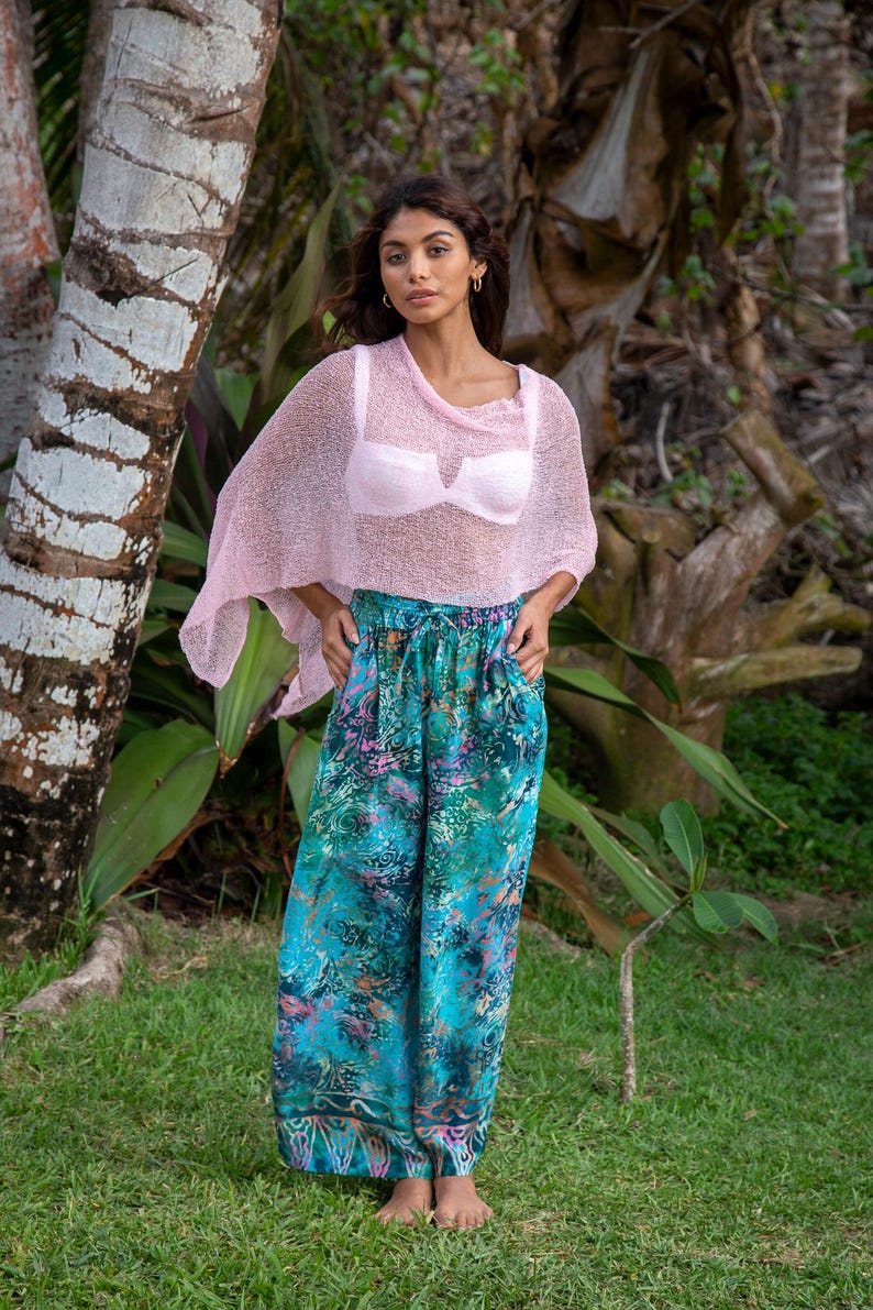 May include: A light pink, sheer, loose-knit poncho worn over a light pink bra, paired with wide-leg trousers featuring a vibrant, multi-coloured abstract pattern in shades of teal, green, blue, and pink with a drawstring waist.