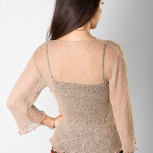 May include: A beige, semi-sheer, long-sleeved top with a textured, crinkled finish, a wide neckline, and ruffled sleeve cuffs and hem, worn over a black camisole, paired with a black skirt featuring a red, yellow, and white floral pattern.