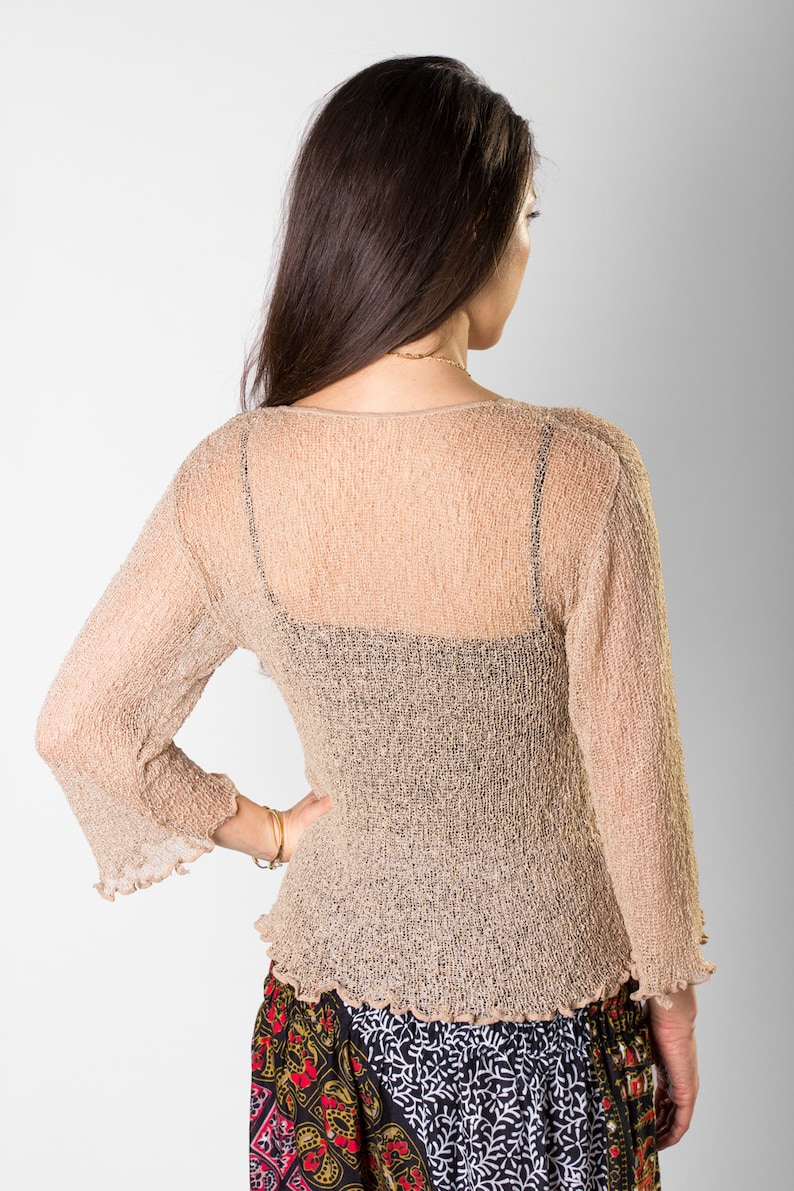May include: A beige, semi-sheer, long-sleeved top with a textured, crinkled finish, a wide neckline, and ruffled sleeve cuffs and hem, worn over a black camisole, paired with a black skirt featuring a red, yellow, and white floral pattern.