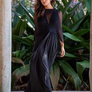 May include: A black sleeveless maxi dress featuring a draped skirt and a sheer, textured black overlay top with a high neckline. The dress is paired with beige wedge sandals with ankle straps, creating a cohesive and layered appearance.