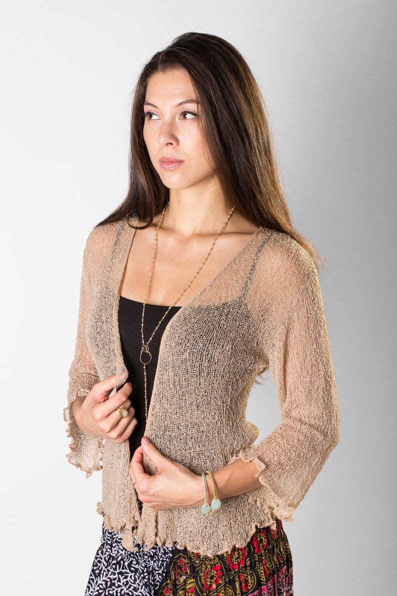 May include: A beige open-front cardigan with a sheer, loose-knit texture, three-quarter length sleeves featuring ruffled edges, and a V-neckline. The garment is layered over a black tank top and paired with a patterned skirt featuring red and white.