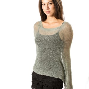 Op de afbeelding: Een saliegroene, losgebreide, semi-transparante poncho met een wijde halslijn, gedragen over een zwart hemdje en zwarte shorts met een geschulpte, gehaakte kanten zoom. De lichtgewicht, textuurrijke gebreide stof valt asymmetrisch over de schouders.