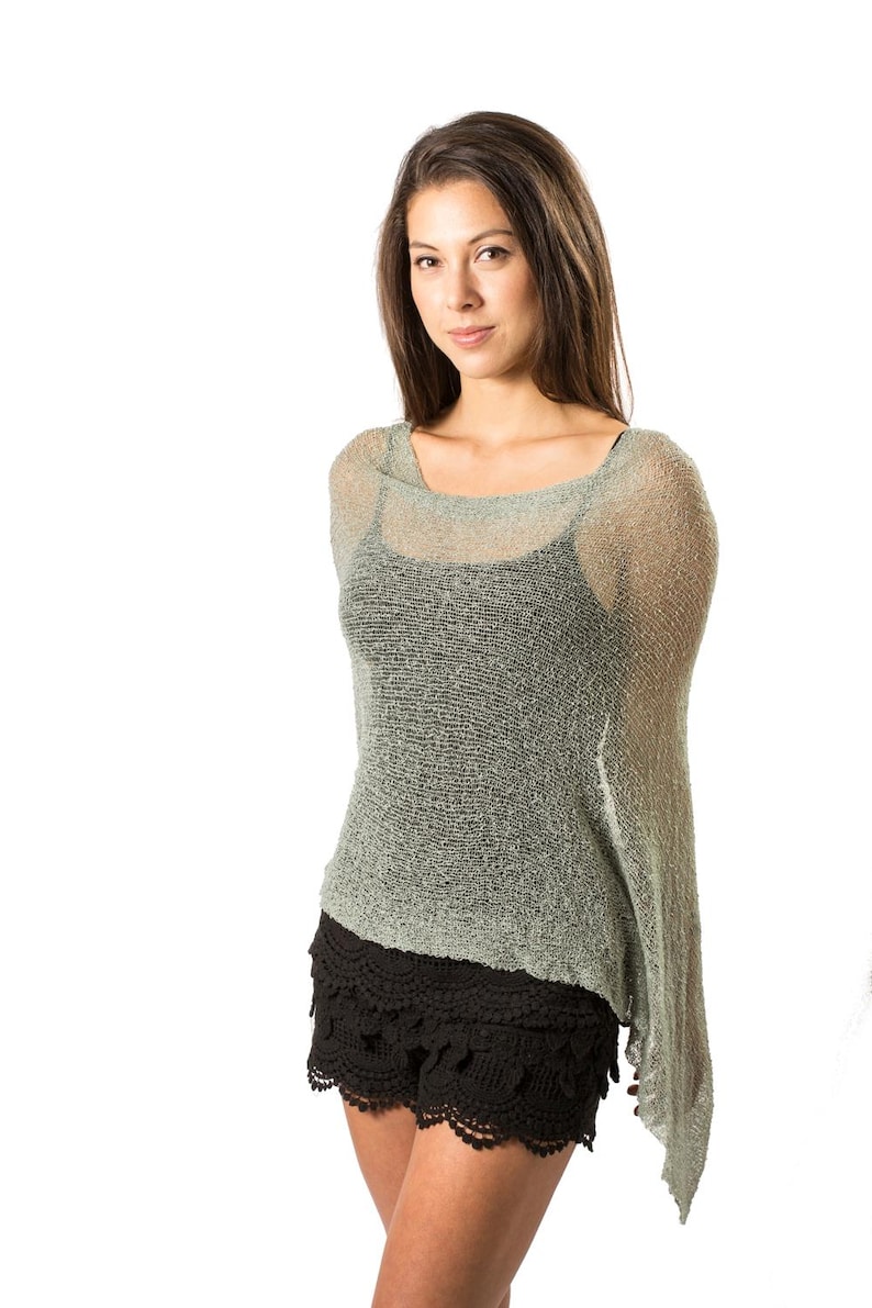 Op de afbeelding: Een saliegroene, losgebreide, semi-transparante poncho met een wijde halslijn, gedragen over een zwart hemdje en zwarte shorts met een geschulpte, gehaakte kanten zoom. De lichtgewicht, textuurrijke gebreide stof valt asymmetrisch over de schouders.