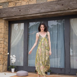Puede incluir: Una mujer con un vestido maxi de estampado floral verde y amarillo con escote en V y tirantes finos se encuentra frente a una pared de piedra y puertas de cristal. Est&aacute; caminando hacia la c&aacute;mara.