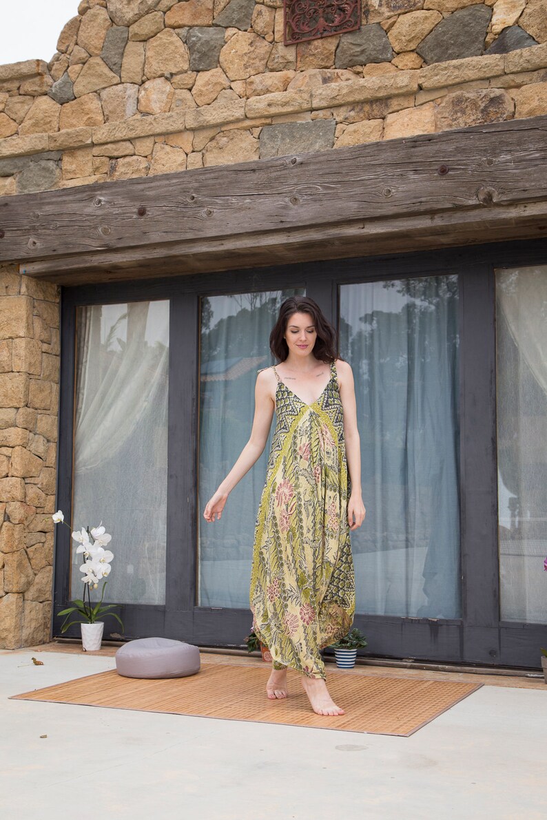 Puede incluir: Una mujer con un vestido maxi de estampado floral verde y amarillo con escote en V y tirantes finos se encuentra frente a una pared de piedra y puertas de cristal. Est&aacute; caminando hacia la c&aacute;mara.