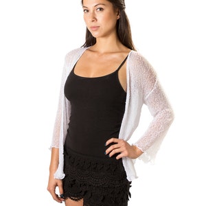 Puede incluir: Un c&aacute;rdigan blanco abierto por delante con una textura de punto fino y transparente y mangas tres cuartos, combinado con una camiseta de tirantes negra y pantalones cortos de encaje negro con dobladillo festoneado y detalles de ganchillo.