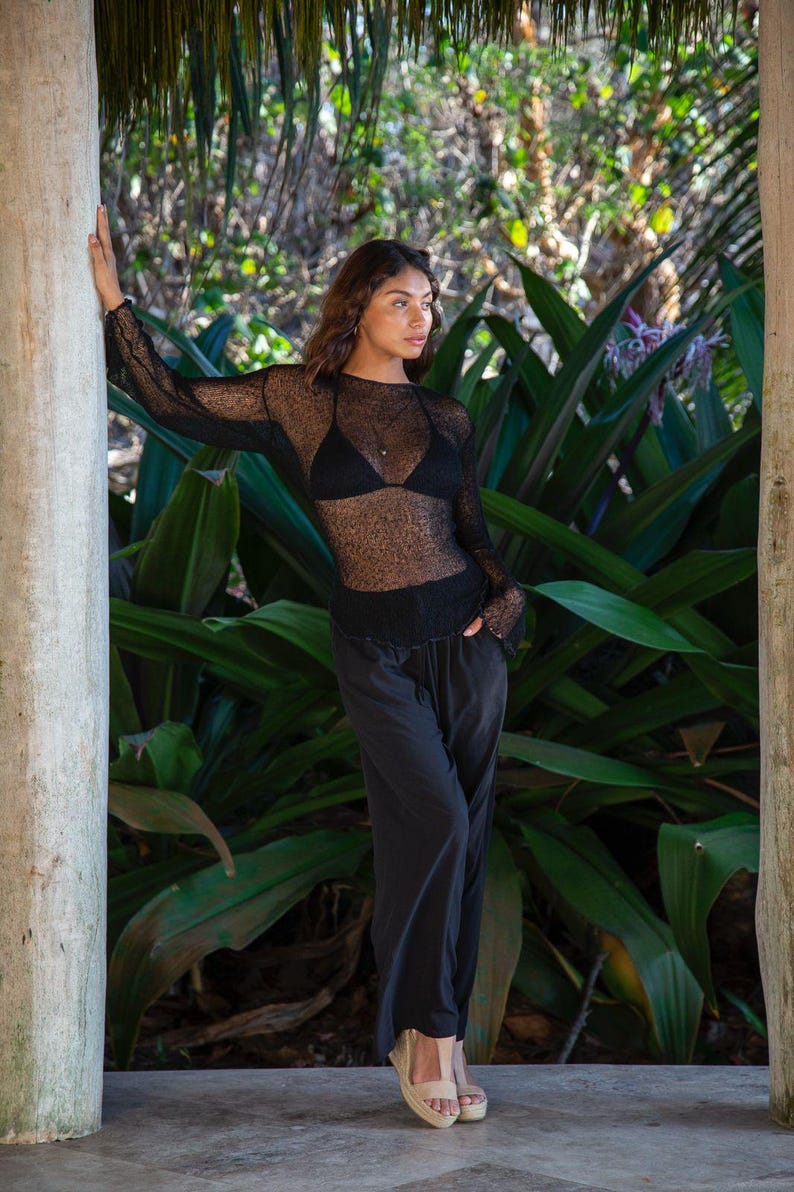 Puede incluir: Una blusa de manga larga de malla negra transparente sobre un top de bikini negro, combinada con pantalones negros holgados y sandalias de cu&ntilde;a beige. El conjunto presenta una textura ligera y semitransparente en la parte superior.