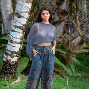 May include: A sheer, slate blue, short-sleeved knit top worn over a light grey bralette, paired with loose-fitting, wide-leg trousers featuring a dark blue base with a multi-coloured, vertical, dash-like pattern and a drawstring waistband.