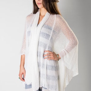 May include: A white crochet poncho with a loose, flowing fit. The poncho is worn over a striped shirt and paired with a white hat.