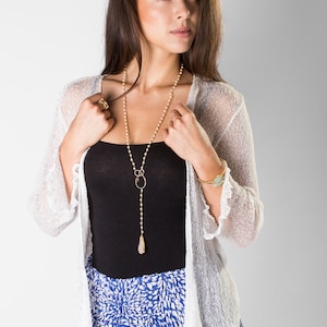 May include: A long necklace featuring small white beads, a gold-toned wire loop, and a teardrop-shaped pendant. The necklace is worn over a black tank top, a white open-front knit cardigan with ruffled edges, and a blue and white patterned skirt.