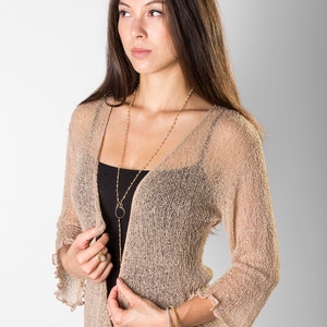 May include: A beige open-front cardigan with a sheer, loose-knit texture and three-quarter length ruffled sleeves. The garment is worn over a black tank top and paired with a patterned skirt featuring black, white, red, and yellow floral motifs.