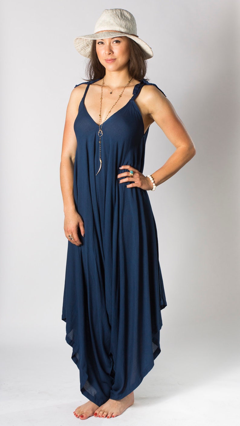 May include: A woman wearing a navy blue jumpsuit with a white hat and a gold necklace. The jumpsuit has a loose, flowing fit and is perfect for a casual summer outfit.