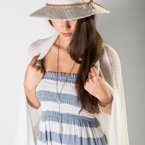 May include: A woman wearing a white crochet cardigan, a blue and white striped dress, and a white woven hat with a brown leather band. She is wearing a silver chain necklace with a turquoise pendant and a silver bracelet with a turquoise stone.