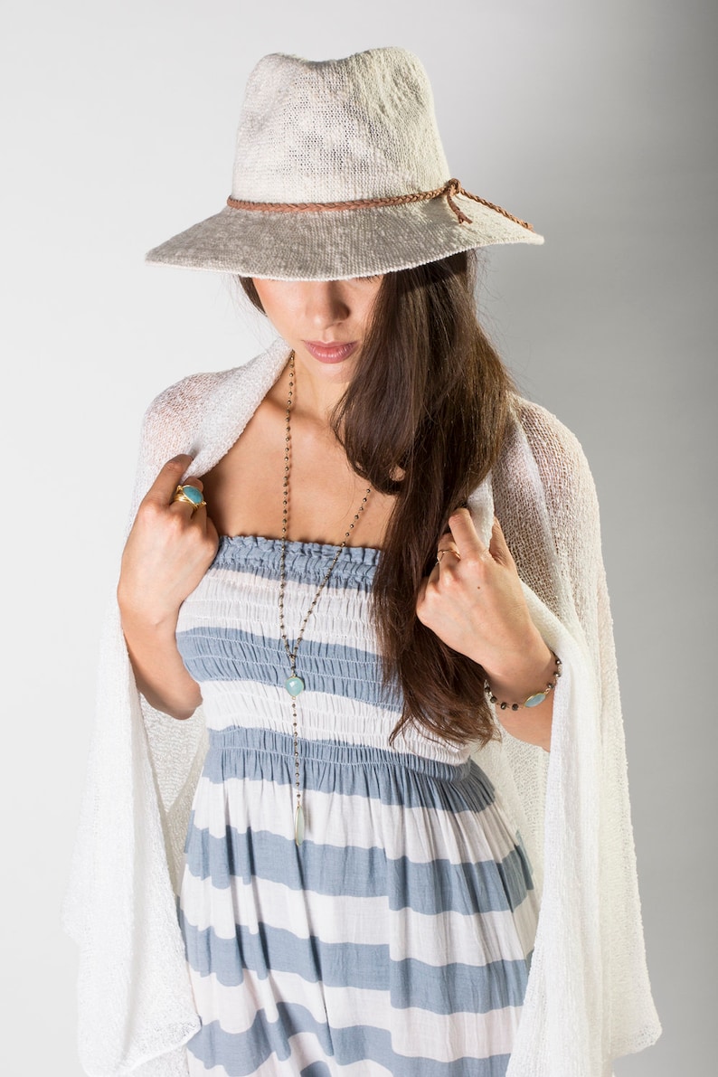 May include: A woman wearing a white crochet cardigan, a blue and white striped dress, and a white woven hat with a brown leather band. She is wearing a silver chain necklace with a turquoise pendant and a silver bracelet with a turquoise stone.
