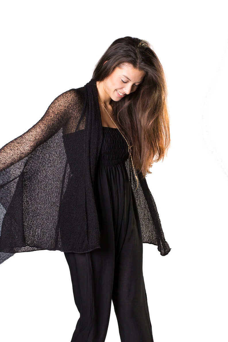 May include: A woman wearing a black sheer cardigan over a black jumpsuit. She is looking down and her long brown hair is flowing.