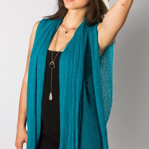 May include: A woman wearing a teal blue knit cardigan with a long gold chain necklace and a white wide-brimmed hat.