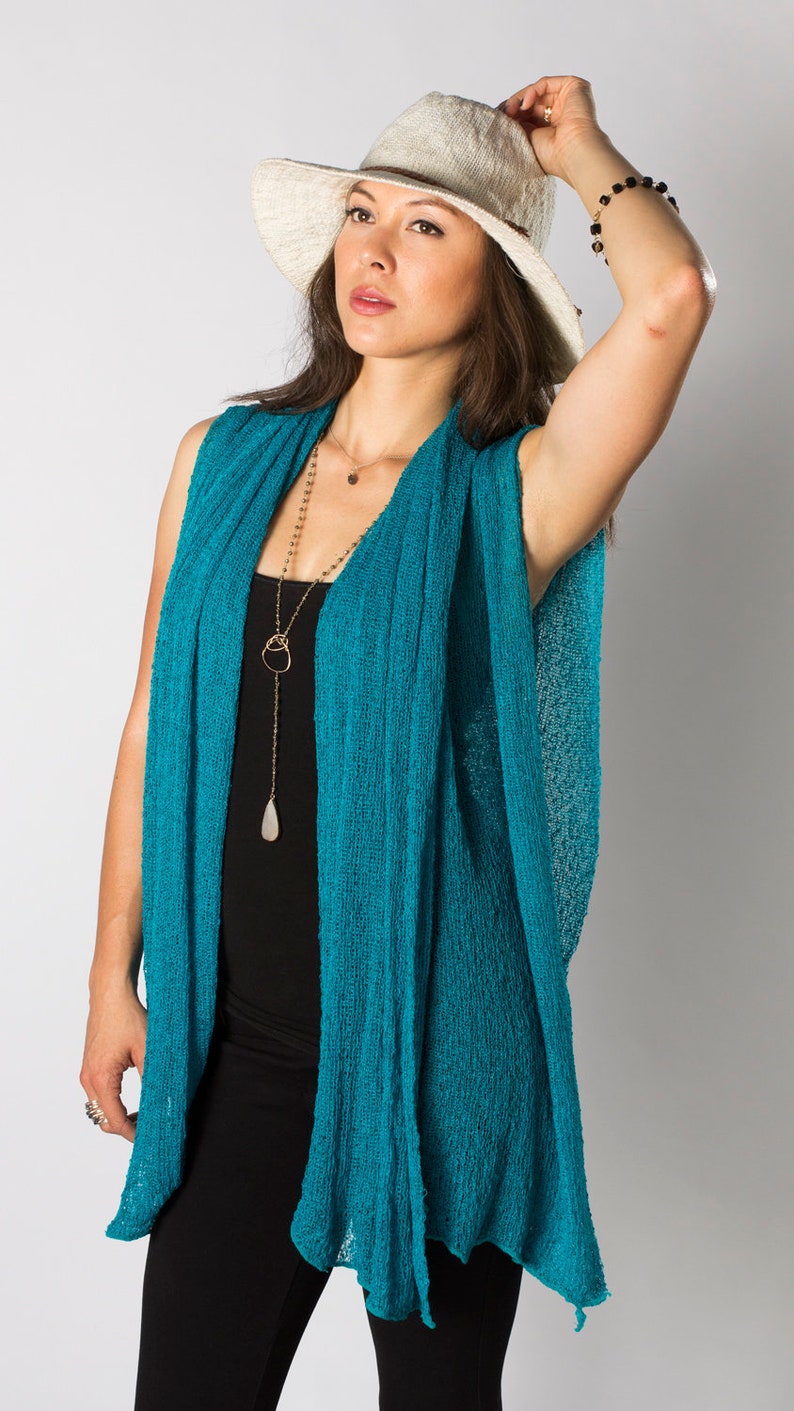 May include: A woman wearing a teal blue knit cardigan with a long gold chain necklace and a white wide-brimmed hat.