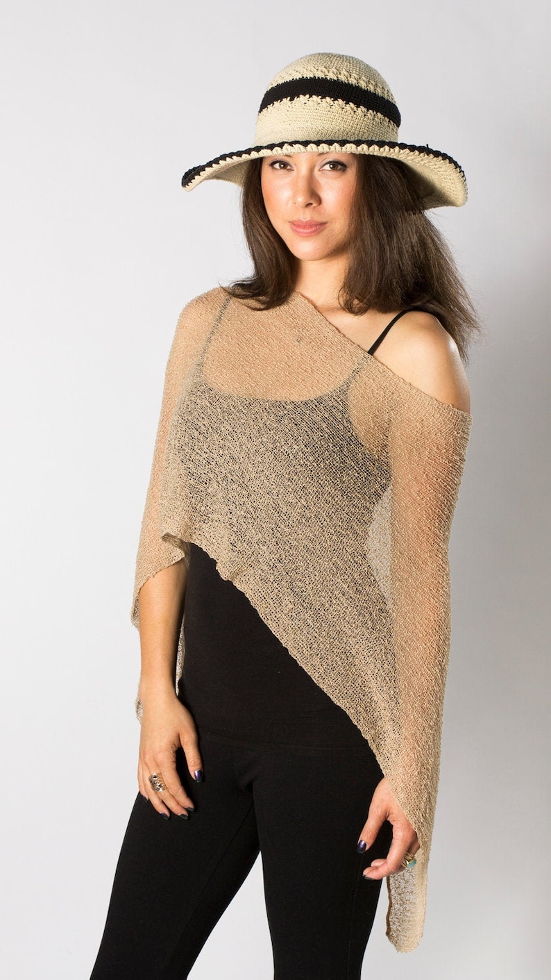 May include: A beige, semi-sheer, asymmetrical knit poncho with a loose, open-weave texture. The garment is worn over a black vest top and black trousers. A beige and black crocheted sun hat with a wide brim is worn on the head.