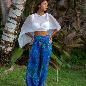 Pu&ograve; includere: Un poncho corto in maglia bianca trasparente indossato sopra un reggiseno bianco, abbinato a pantaloni blu larghi con un vivace motivo astratto multicolore, vita elasticizzata con coulisse e gambe ampie.