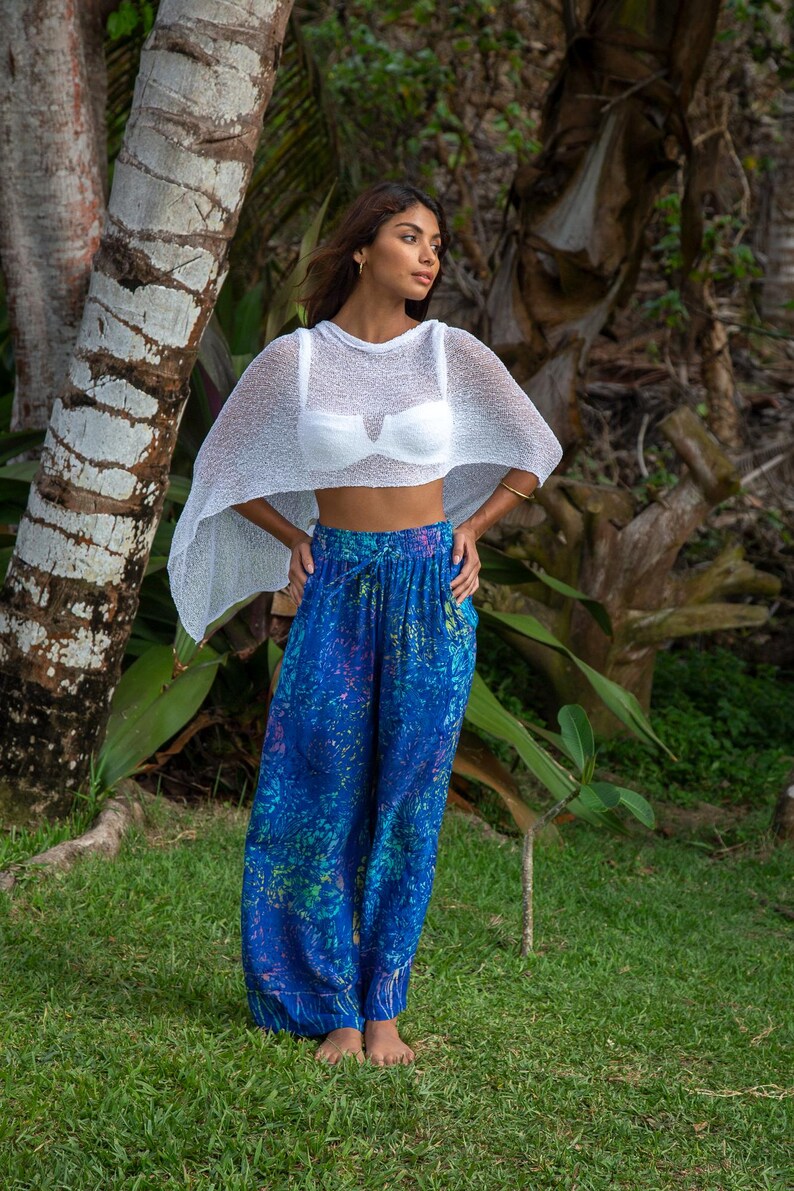 Pu&ograve; includere: Un poncho corto in maglia bianca trasparente indossato sopra un reggiseno bianco, abbinato a pantaloni blu larghi con un vivace motivo astratto multicolore, vita elasticizzata con coulisse e gambe ampie.