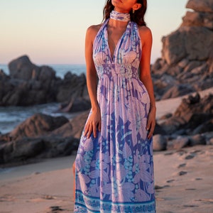 May include: A woman wearing a long, flowing maxi dress with a floral print in shades of purple, blue, and white. The dress has a halter neckline and a fitted bodice. The skirt is full and flowing, with a side slit. The woman is standing on a sandy beach with rocks in the background.