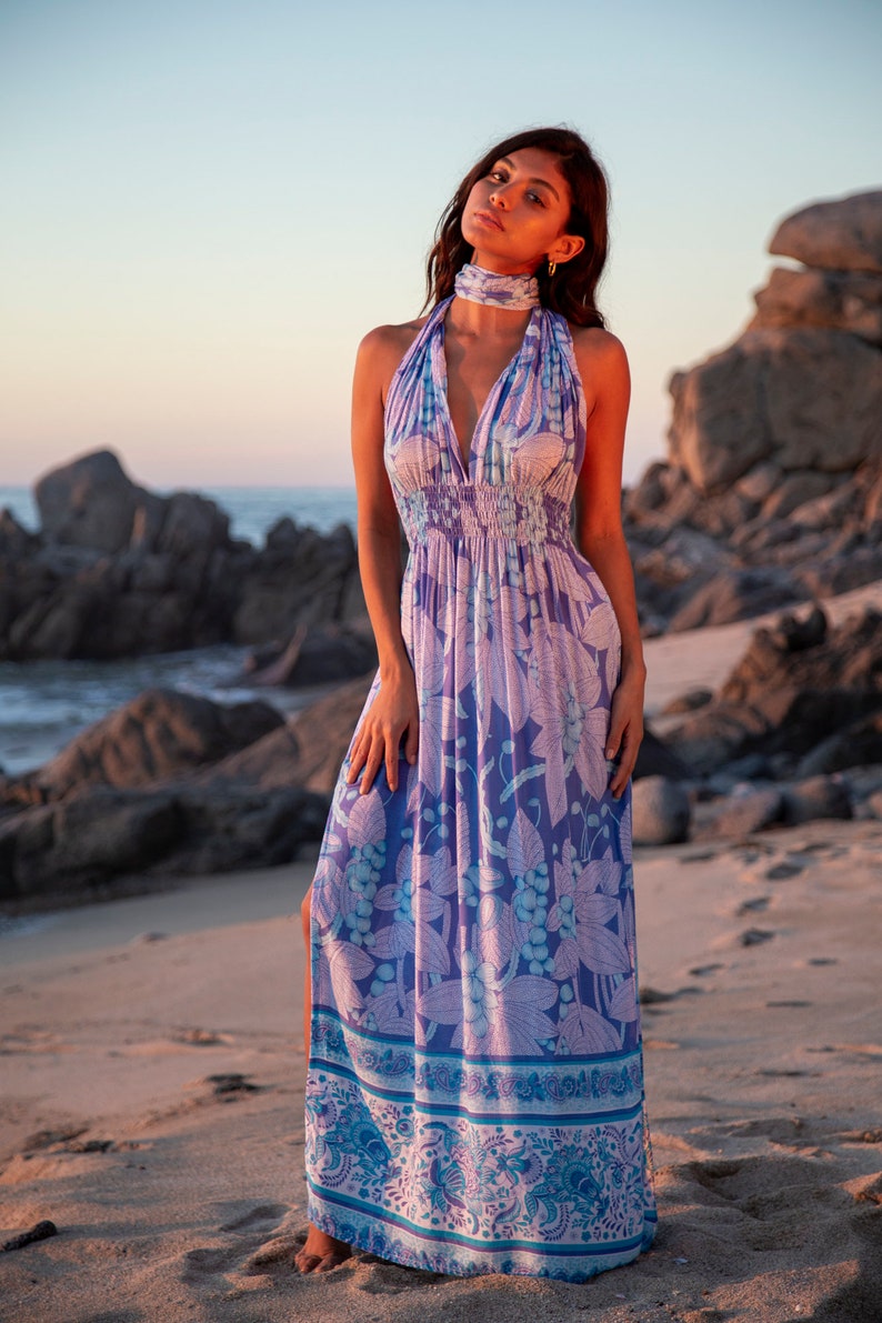 May include: A woman wearing a long, flowing maxi dress with a floral print in shades of purple, blue, and white. The dress has a halter neckline and a fitted bodice. The skirt is full and flowing, with a side slit. The woman is standing on a sandy beach with rocks in the background.