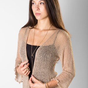 Puede incluir: Un c&aacute;rdigan beige abierto por delante con una textura de punto suelto y transparente, y mangas tres cuartos con volantes. La prenda se lleva sobre una camiseta de tirantes negra y se combina con una falda estampada con motivos florales.