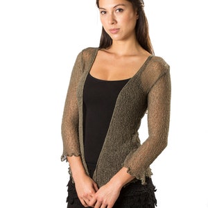 Puede incluir: Un c&aacute;rdigan abierto de color verde oliva con una textura de punto fino y transparente, con mangas tres cuartos y bordes con volantes. La prenda se lleva sobre una camiseta de tirantes negra lisa y pantalones cortos negros con un dobladillo de encaje.