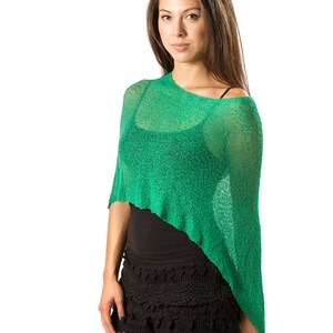 Op de afbeelding: Een felgroene, semi-transparante gebreide poncho met een asymmetrische zoom. Het kledingstuk heeft een losse, open breistructuur en een wijde halslijn die over de schouders valt, gedragen over een zwart hemdje en een zwarte kanten short.