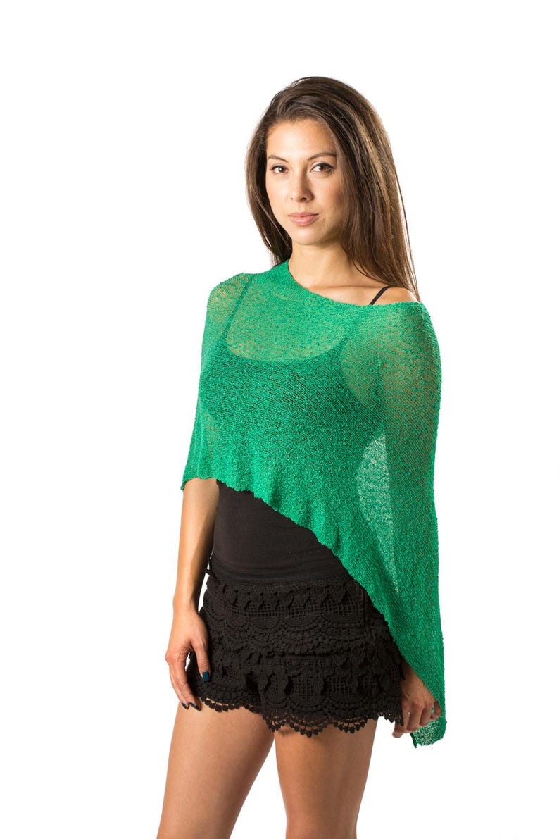 Op de afbeelding: Een felgroene, semi-transparante gebreide poncho met een asymmetrische zoom. Het kledingstuk heeft een losse, open breistructuur en een wijde halslijn die over de schouders valt, gedragen over een zwart hemdje en een zwarte kanten short.