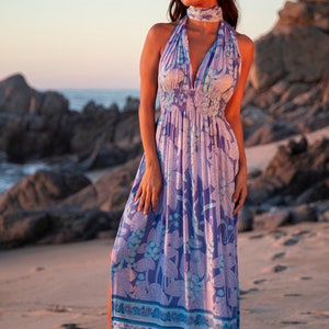 May include: A woman wearing a long, flowing purple and blue floral print maxi dress with a halter neckline and a tie at the neck. The dress has a gathered waist and a side slit. The woman is standing on a sandy beach with rocks in the background.