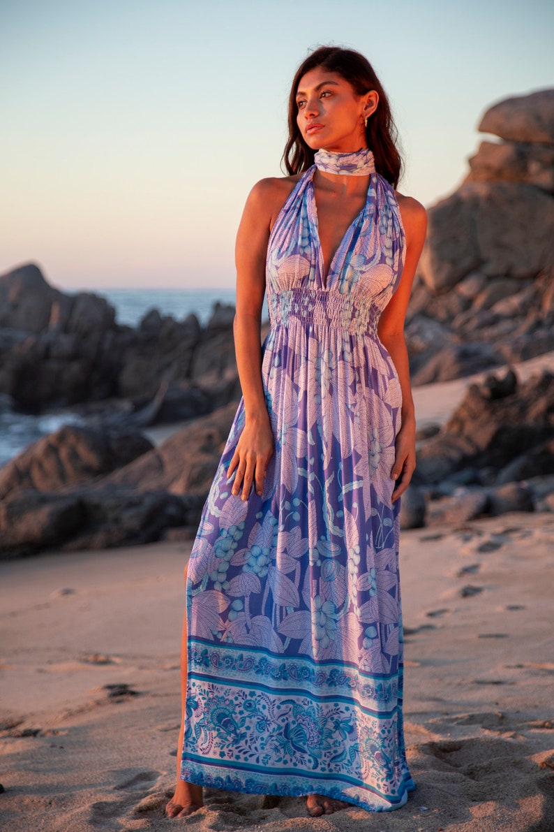 May include: A woman wearing a long, flowing purple and blue floral print maxi dress with a halter neckline and a tie at the neck. The dress has a gathered waist and a side slit. The woman is standing on a sandy beach with rocks in the background.