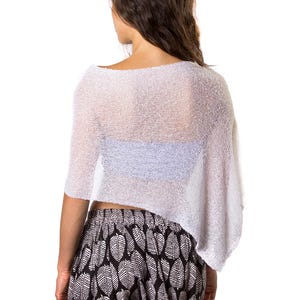 Pu&ograve; includere: Un top a poncho bianco, in maglia larga e semitrasparente con orlo asimmetrico, indossato sopra un capo azzurro, abbinato a pantaloncini neri con motivo a foglie bianche e bordo con pompon bianchi lungo l'orlo inferiore.