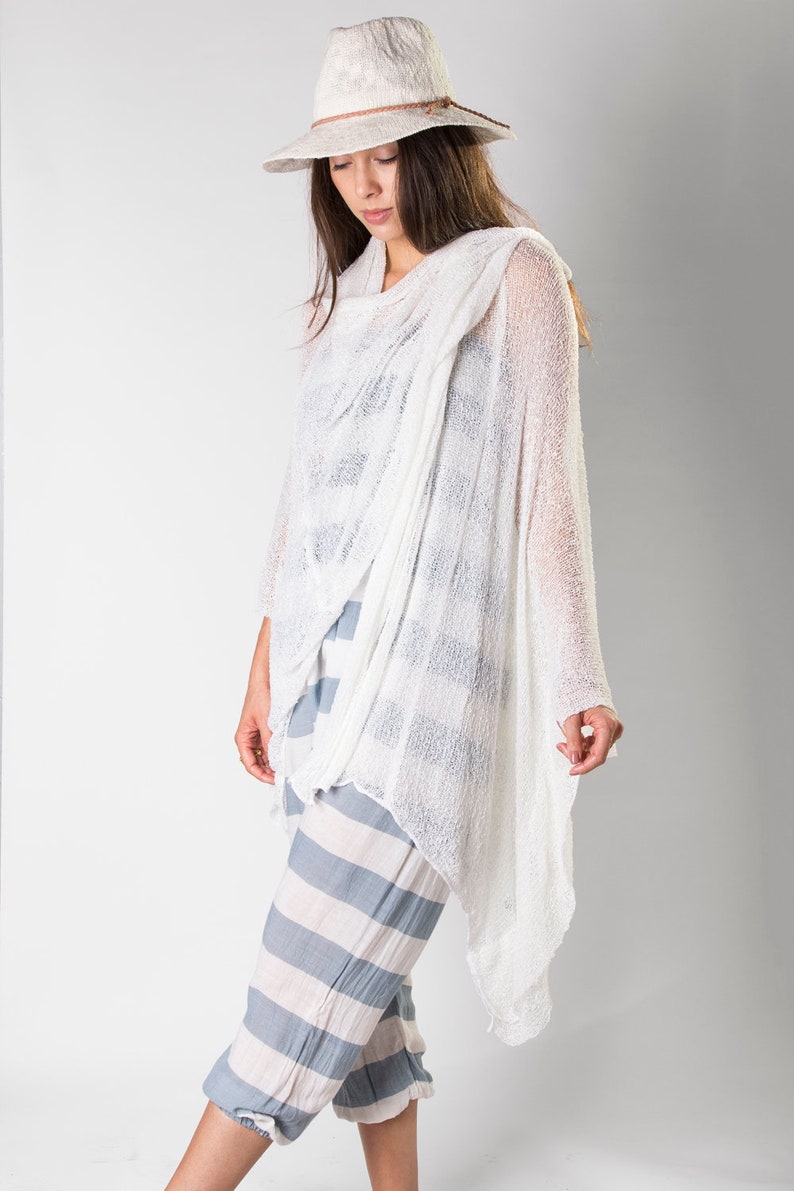 May include: A white crochet top with a loose, draped fit. The top has a wide, open neckline and long sleeves. The model is wearing the top with striped pants and a white hat.