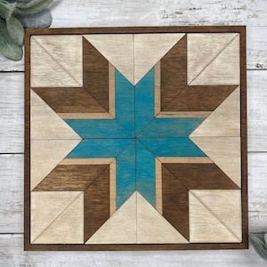 May include: A wooden wall art piece featuring a geometric star design. The star is crafted from light brown and dark brown wood, with a teal blue centre. The piece is framed in a dark brown wood frame.