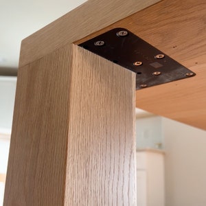 May include: Close-up of a wooden table leg attached to the tabletop with a black metal bracket and screws. The wood grain is visible on the table leg and tabletop.