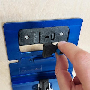 May include: A blue and black woodworking jig with a black plastic lever. The jig has a small hole and a slot for a router bit.