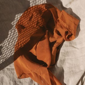 May include: A draped, burnt orange scarf made of a lightweight fabric. The scarf is arranged on a neutral-colored surface, with shadows from a lattice pattern overlaying the fabric. The scarf's folds and texture are highlighted by the sunlight.