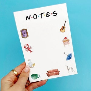 May include: White notepad with the text "NOTES" in black lettering. The notepad has various illustrations from the TV show Friends, including a purple picture frame, a red lobster, a white duck, a yellow chick, a red couch, a blue umbrella, a guitar, and a turkey wearing sunglasses and a red hat.
