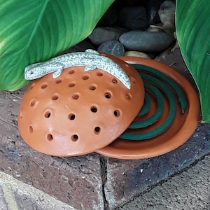 以下が含まれることがあります： テラコッタ製の香炉。上部にセラミック製のトカゲが乗っており、内部には緑色の渦巻き状の線香が入っています。香炉は丸い形をしていて、通気口の穴が開いています。