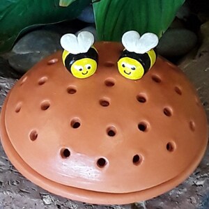 May include: Two small ceramic bee figurines with yellow and black paint and white wings are perched on a terracotta pot with drainage holes. The pot is brown and has a rounded shape.