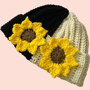 May include: A handmade crochet beanie in black, cream, and white, adorned with two yellow and brown sunflower appliqués. The hat is made of soft yarn and features a ribbed texture. A stylish and unique accessory.