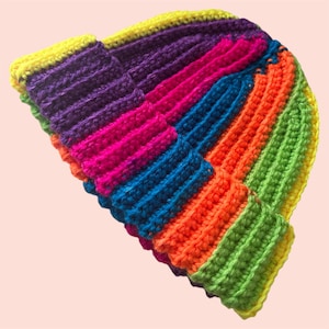 May include: A handmade, colorful beanie hat. The hat is crocheted with vibrant rainbow colors, including yellow, purple, pink, blue, orange, and green. The hat has a ribbed texture and a folded brim. The hat is a stylish accessory.